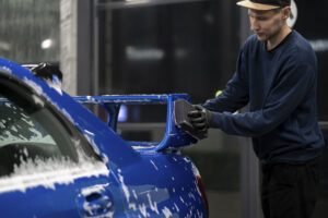 Eine Person reinigt ein blaues Auto mit einer Bürste, um Schnee und Eis zu entfernen. Der Fahrzeugpflege Winter Tipps Ratgeber empfiehlt diese Methode, um das Fahrzeug winterfit zu halten.
