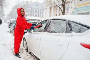 Eine Person in roter Winterkleidung entfernt Schnee von der Windschutzscheibe eines weißen Autos. Ein wichtiger Tipp aus dem Fahrzeugpflege Winter Ratgeber für klare Sicht im Winter.