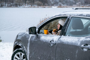Eine Person sitzt in einem verschneiten Auto mit einer Tasse Kaffee, während Schnee das Fahrzeug bedeckt. Fahrzeugpflege Winter Tipps helfen, das Auto für kalte Tage vorzubereiten.