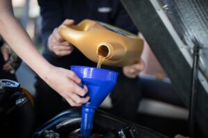 Zwei Personen füllen Motoröl mit einem blauen Trichter in den Motorraum eines Autos. Der Ölverlust Auto erkennen Ratgeber empfiehlt, nach einem Ölverlust den Ölstand wieder aufzufüllen.
