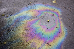 Ein bunter Ölfleck mit Regenbogeneffekt auf einer nassen Straße, ein klares Zeichen für Ölverlust. Der Ölverlust Auto erkennen Ratgeber hilft, solche Anzeichen frühzeitig zu identifizieren.
