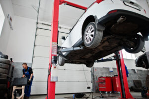 Ein weißes Auto steht auf einer Hebebühne in einer Werkstatt, während ein Mechaniker arbeitet. Der Reifenrotation Häufigkeit Tipps Ratgeber empfiehlt regelmäßige Checks in der Werkstatt.
